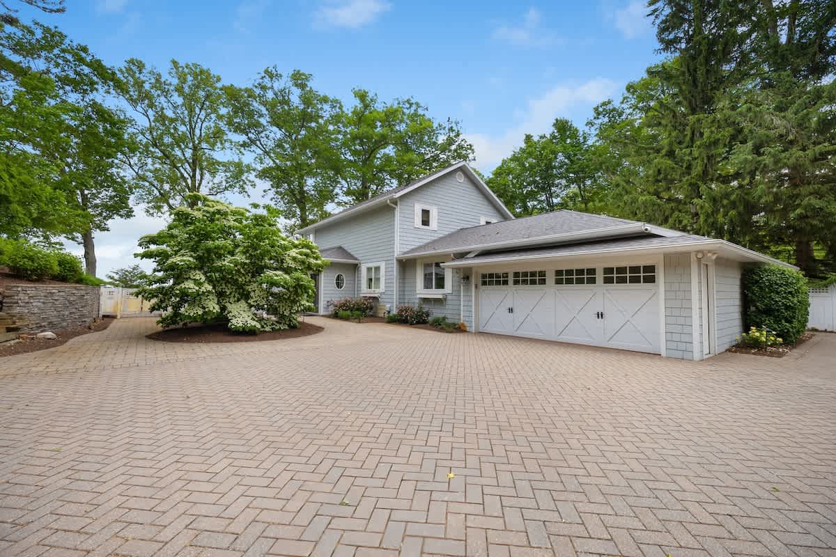 Front exterior of the light blue house with a large paved herringbone driveway, an attached two-car garage, and mature landscaping including a blooming white bush.