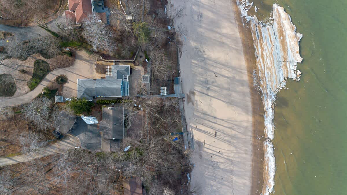 Overhead aerial view showing the property nestled among trees next to a wide sandy beach and a large body of water, with some ice formations along the shoreline.