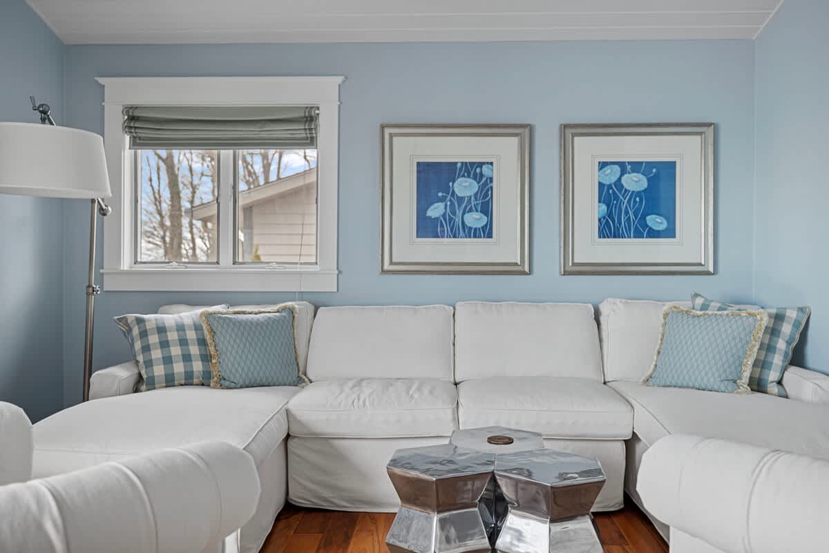 Light blue living room featuring a white sectional sofa, two silver geometric coffee tables, and two framed blue botanical prints on the wall. A window with a gray Roman shade is visible on the left, complementing the light blue walls and hardwood floors.