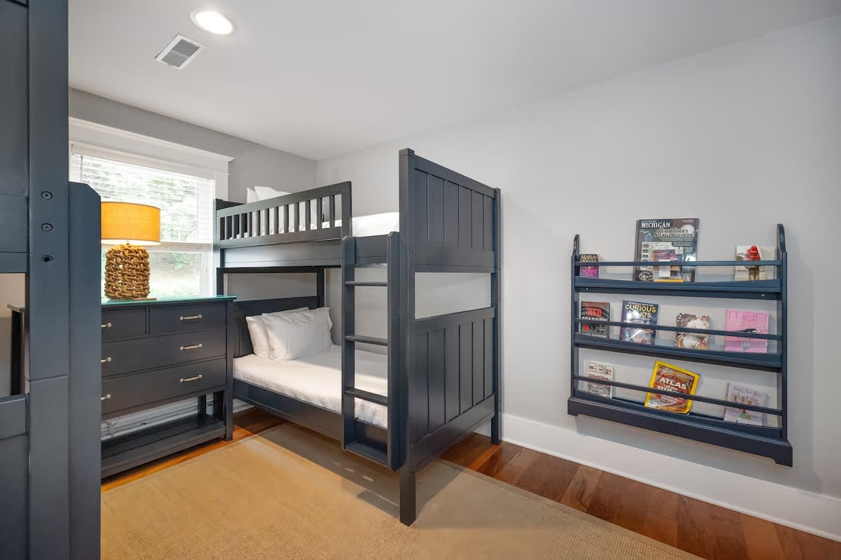 Kids' bedroom with a dark blue wooden bunk bed on the right and a matching dresser with a woven lamp under a window on the left. A wall-mounted bookshelf filled with books is visible on the far right, all set on a light brown rug.