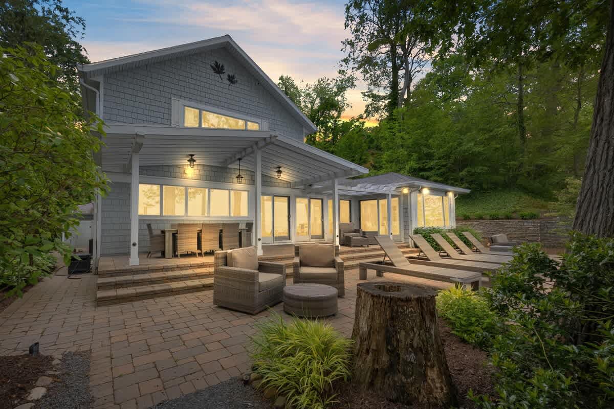 Rear exterior of a light gray shingle-sided house with a covered patio featuring outdoor seating and lounge chairs, overlooking a large body of water at dusk.