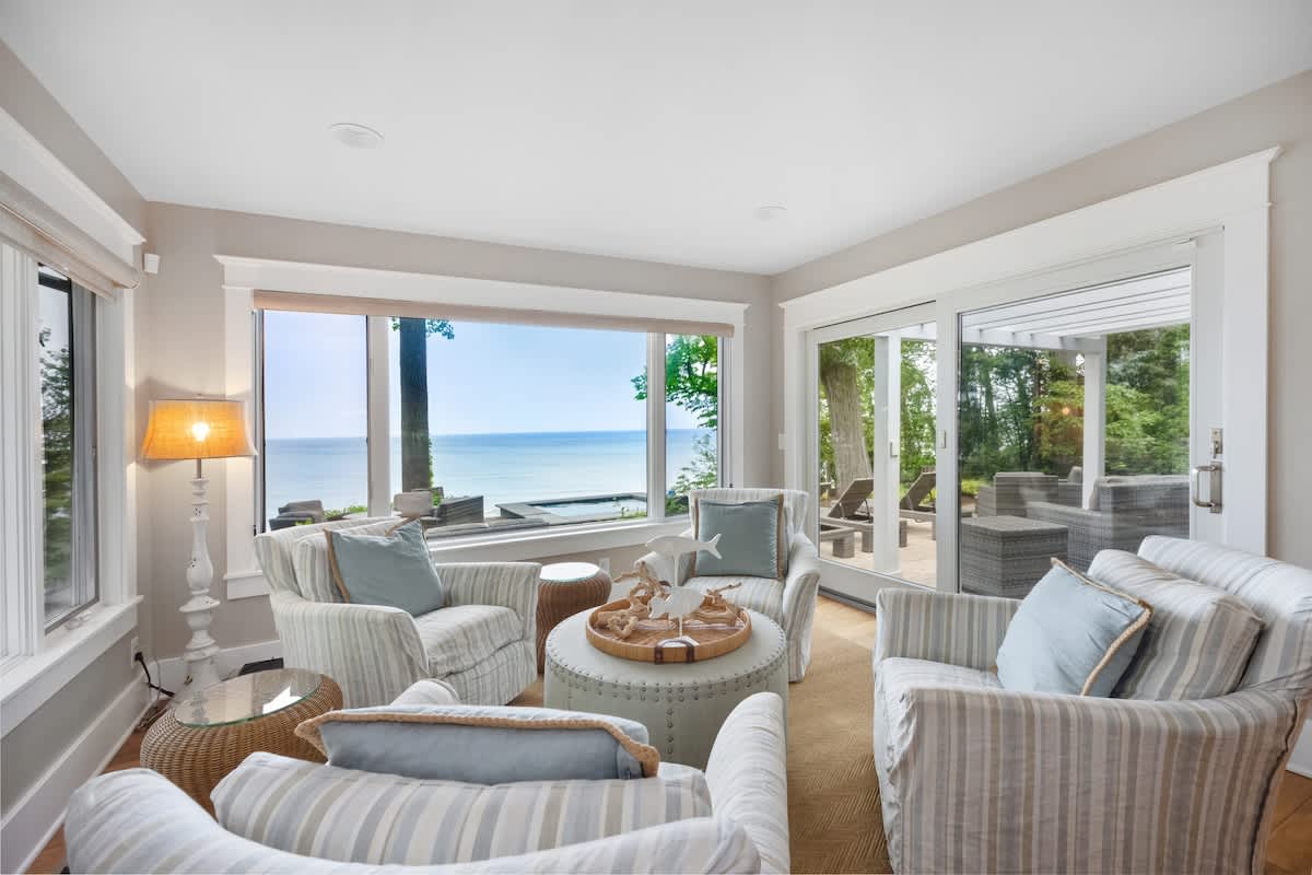 A bright living room features four striped armchairs arranged around a round upholstered ottoman, with large windows and sliding glass doors offering expansive views of the lake and outdoor patio. A decorative lamp stands in the corner.