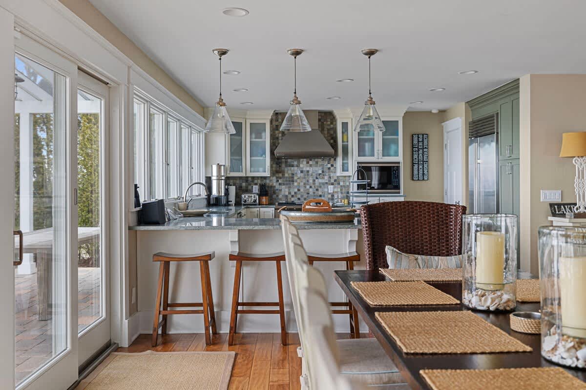 The dining area, with a dark wood table and wicker chairs, transitions into an open kitchen featuring a breakfast bar with stools, light cabinetry, stainless steel appliances, and large windows.
