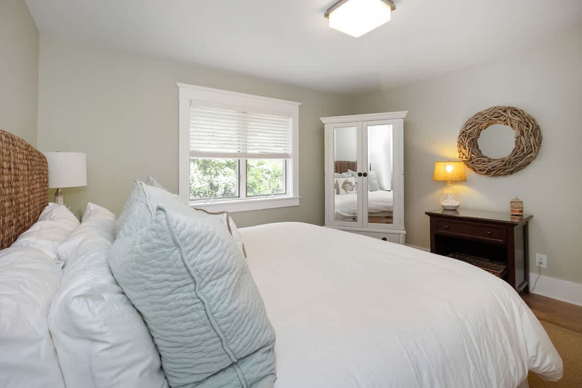 Cozy bedroom with a king-sized bed, woven headboard, and white bedding. A dark wood nightstand with a lamp and a large woven wreath on the wall are visible on the right, next to a white armoire and a window with blinds.