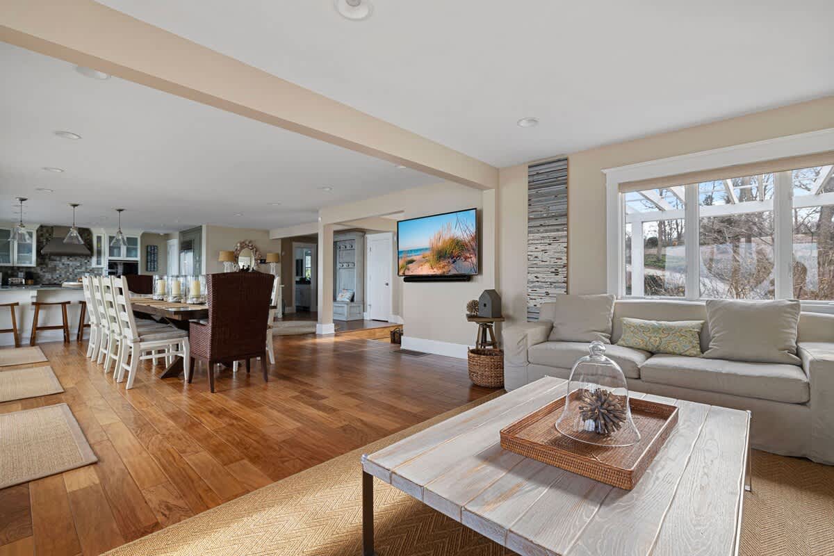 An open-concept living space features light-colored walls, hardwood floors, a comfortable sofa, a light wood coffee table, and a wall-mounted TV, flowing into the dining area and kitchen beyond.