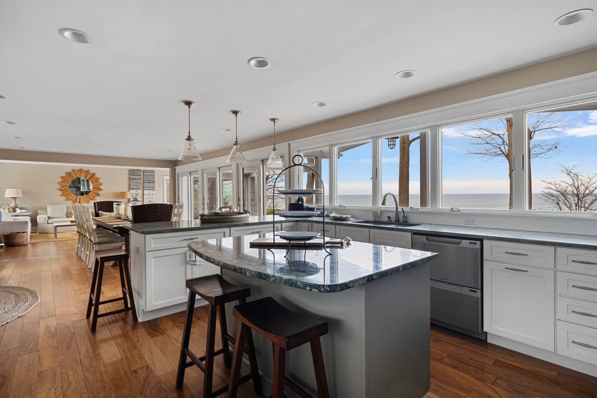 Spacious kitchen with white cabinetry, light gray granite countertops, and a long window providing a panoramic lake view. A central island with bar stools offers additional seating, opening to a living area.