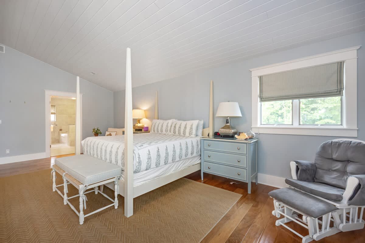 Another view of the light blue bedroom, highlighting the white four-poster bed, a light blue dresser with a lamp, and a gray glider chair. A window with a striped Roman shade offers natural light.