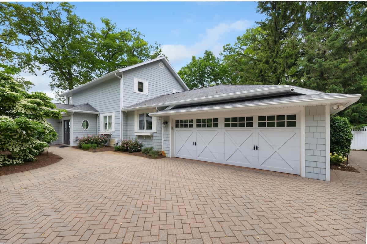 The front exterior of the light blue shingle-sided house features a large white two-car garage with cross-brace doors and a herringbone brick paver driveway, surrounded by lush green trees and shrubs.