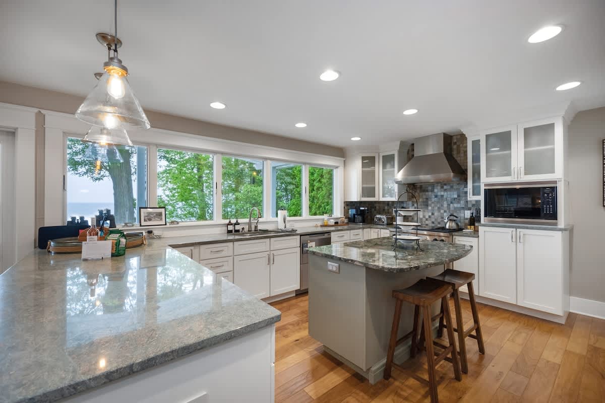 Bright, modern kitchen with white shaker cabinets, light gray granite countertops, and a large window offering a lake view. It features a central island with seating, stainless steel appliances, and hardwood floors.