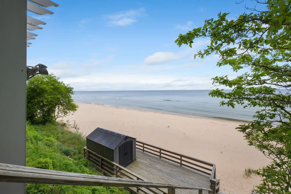 A wooden walkway with railings descends towards a wide sandy beach and the vast ocean, with a small dark shed positioned near the water's edge. Lush green foliage frames the path.