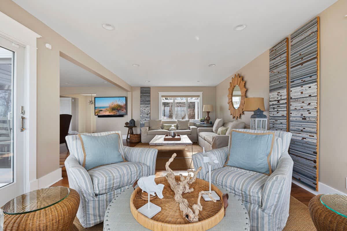 A cozy living area features two striped armchairs with light blue pillows facing a round ottoman with coastal decor, with a view into a larger living space beyond, all on hardwood floors.