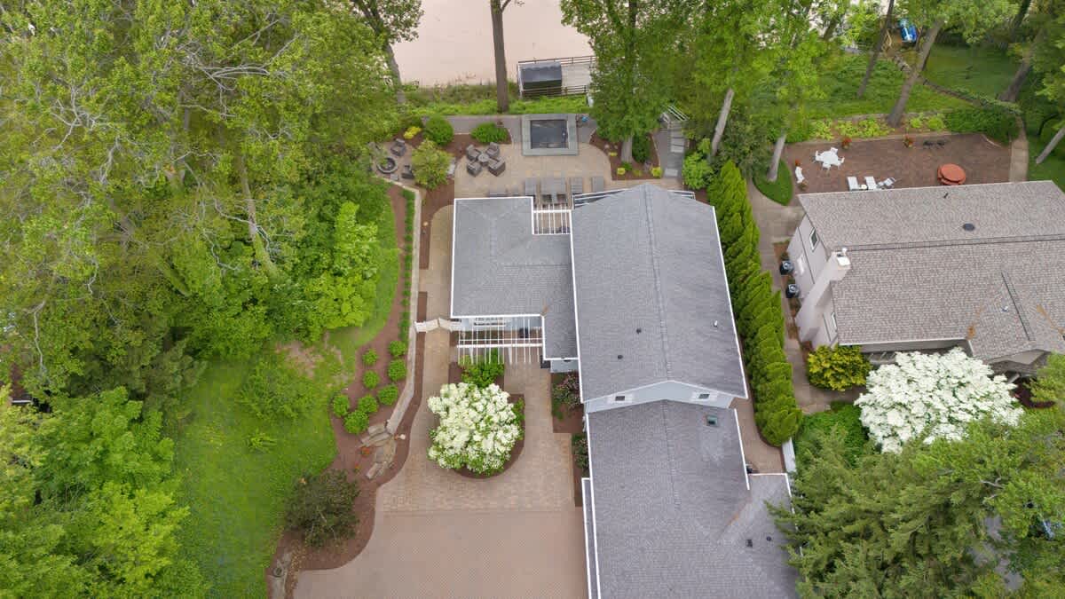 Closer aerial view of the property, highlighting the house's roofline, the paved patio with lounge chairs, and the fire pit area nestled among the trees by the lake.
