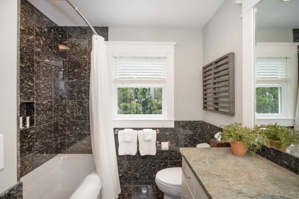 View of the bathroom with dark granite tile walls and floor, showcasing the shower/tub combo with a white curtain and a window with white blinds. A toilet is visible next to the vanity.