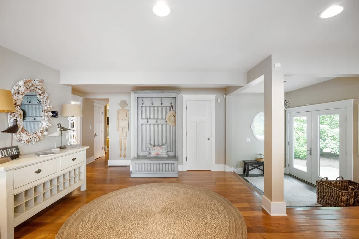 A welcoming foyer features warm hardwood floors, a round jute rug, a white console table with coastal decor, and a built-in gray bench with coat hooks, leading to an entryway with double glass doors.