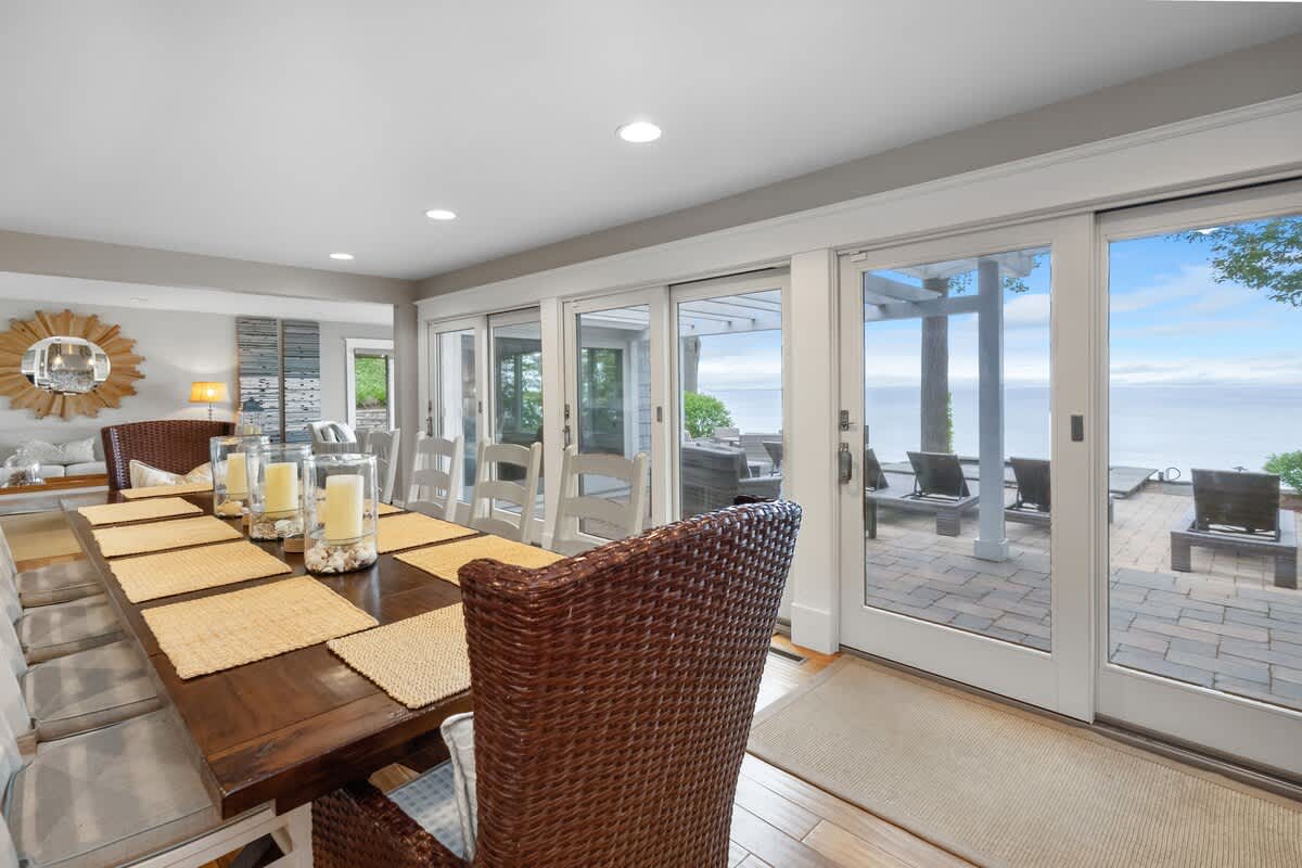 A spacious dining room features a long dark wood table with wicker chairs and white chairs, set on hardwood floors, with multiple sliding glass doors offering expansive views of the lake and leading to an outdoor patio.