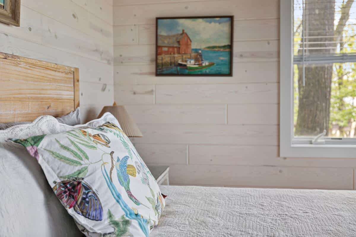 Close-up of a bedroom with light wood paneled walls, showing a white bed with a colorful floral pillow and a framed painting of a coastal scene above the headboard.