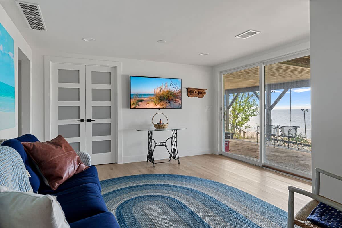 Modern living area with light wood flooring and white walls, featuring a wall-mounted TV above a small glass-top table. Large sliding glass doors provide expansive views of the beach and water.