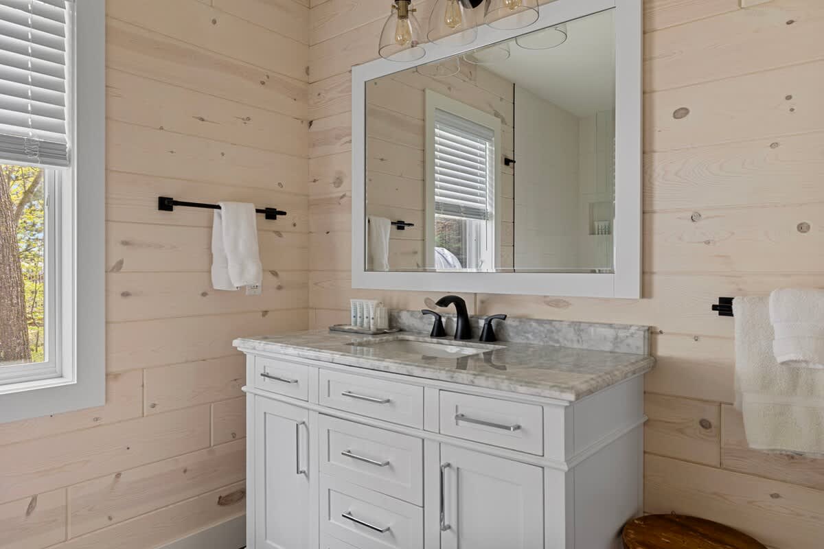 A bright bathroom with light wood paneled walls, a white vanity with a marble countertop, and a large mirror. A window with blinds offers natural light.
