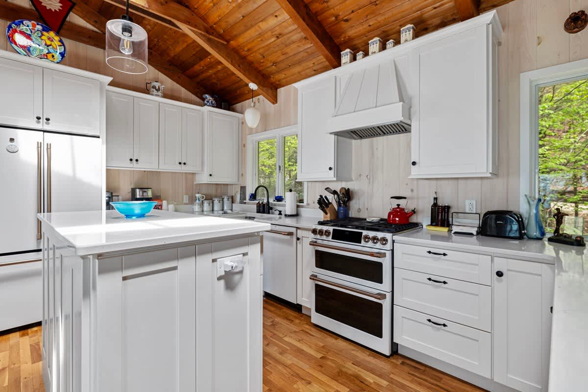 Bright white kitchen featuring a double oven range, white cabinets, and light countertops, with light wood floors and a vaulted wood ceiling. A large window above the sink offers a view of lush greenery outside.