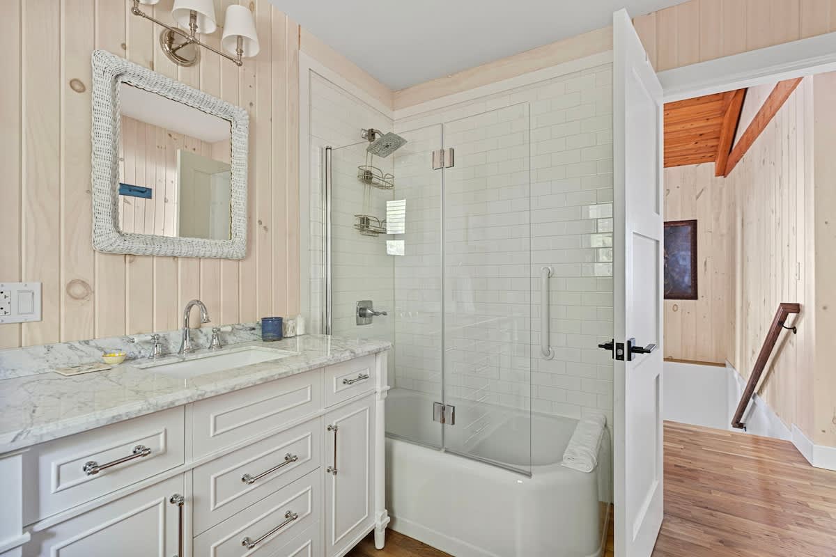 Light and airy bathroom featuring a white marble-topped vanity with a single sink, white shaker cabinets, and a glass-enclosed shower-over-tub with white subway tiles. The walls are paneled with light wood.