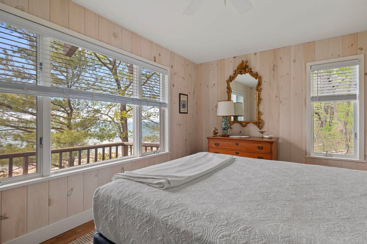 A bedroom with light wood paneled walls and a white bed, featuring a large window that provides a scenic view of trees and the distant ocean.