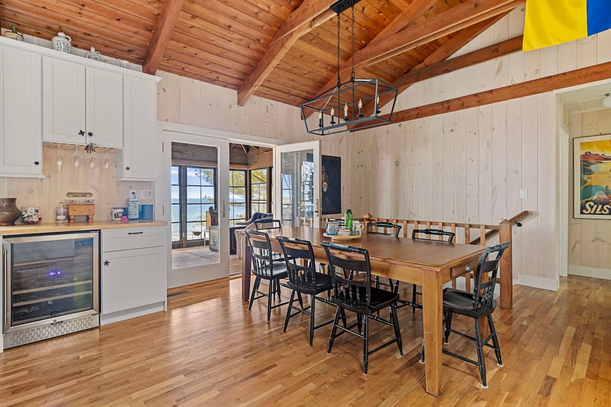 Dining area with a long wooden table and black chairs, featuring light wood floors and a vaulted wood ceiling. A white wet bar with a beverage fridge is visible on the left, and glass doors lead to an outdoor space.