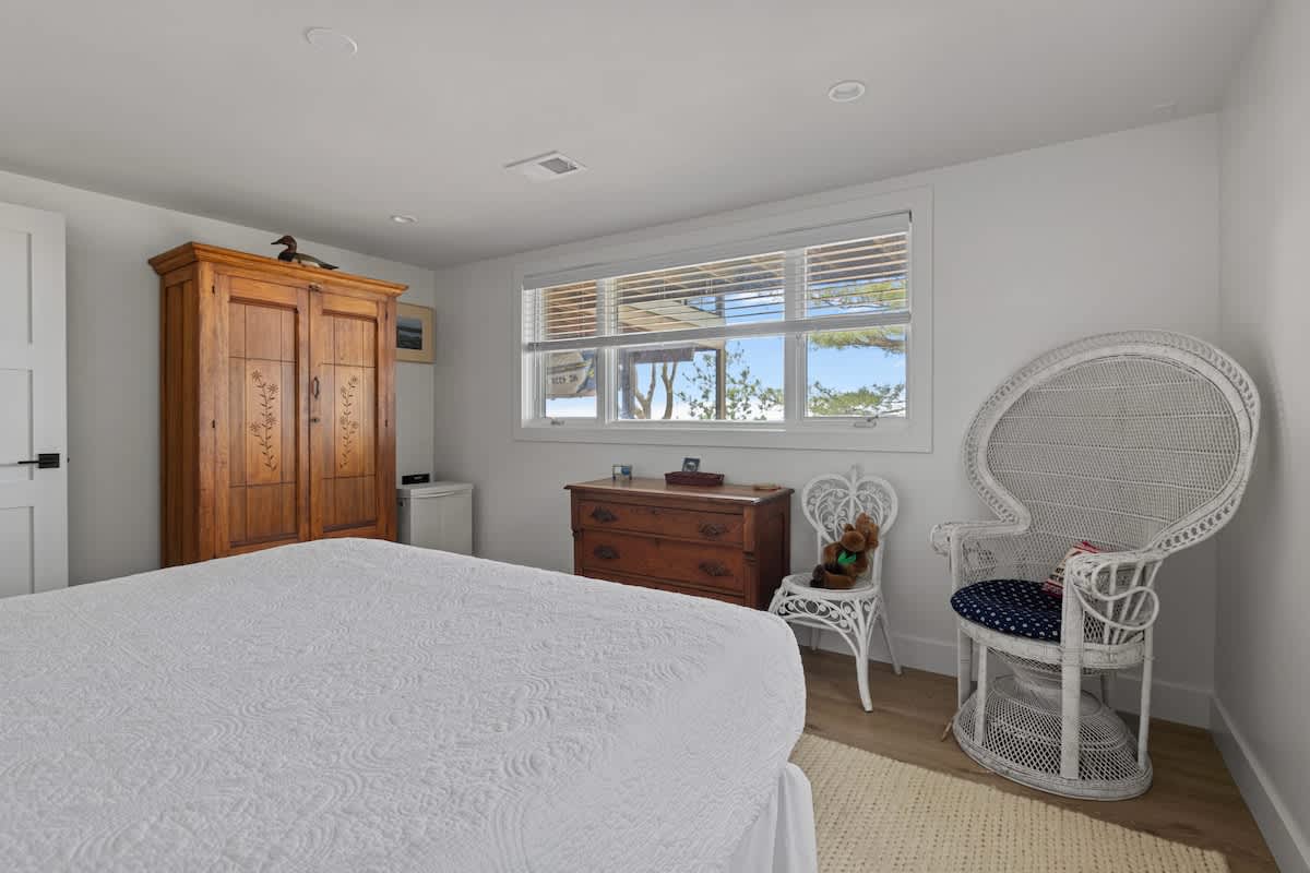White-walled bedroom with light wood flooring, featuring a white bed, a large wooden armoire, and a vintage wooden dresser. A white wicker chair and a large window with blinds are also visible.