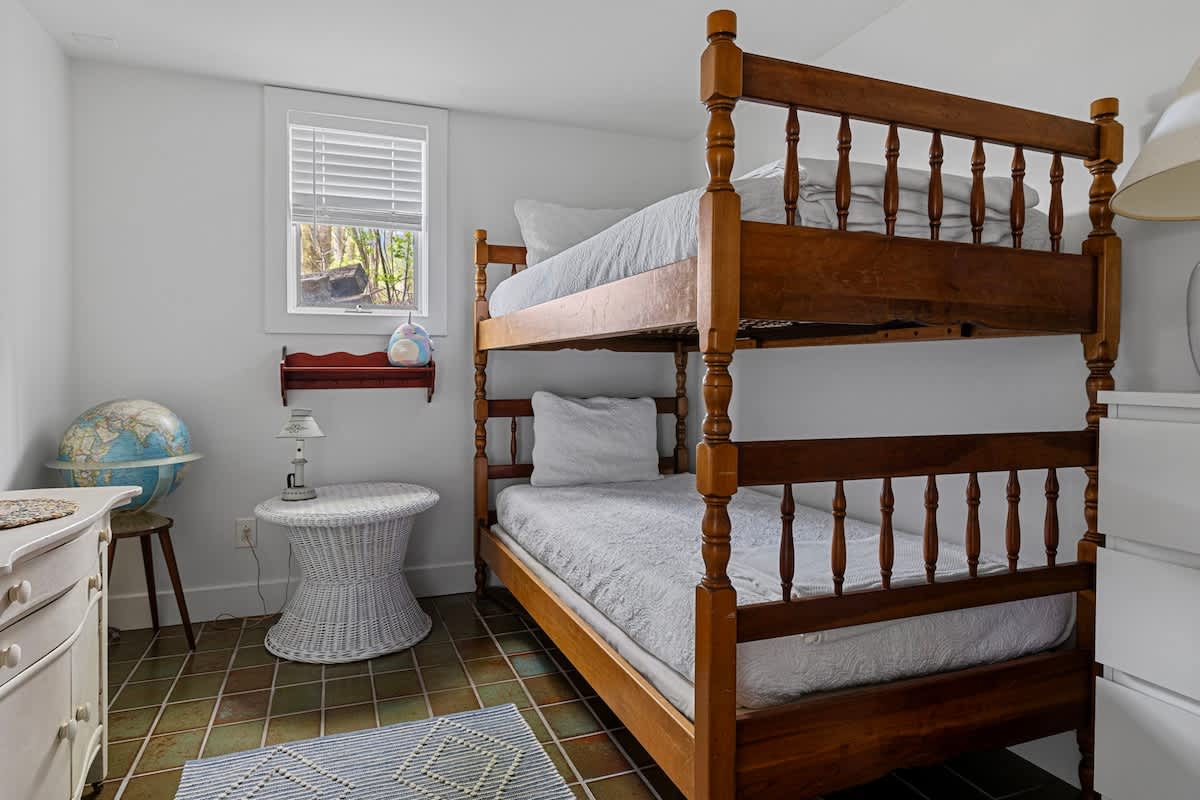 Children's bedroom with white walls and tiled flooring, featuring a wooden bunk bed with white bedding. A small window with blinds, a white wicker side table, and a globe are also visible.