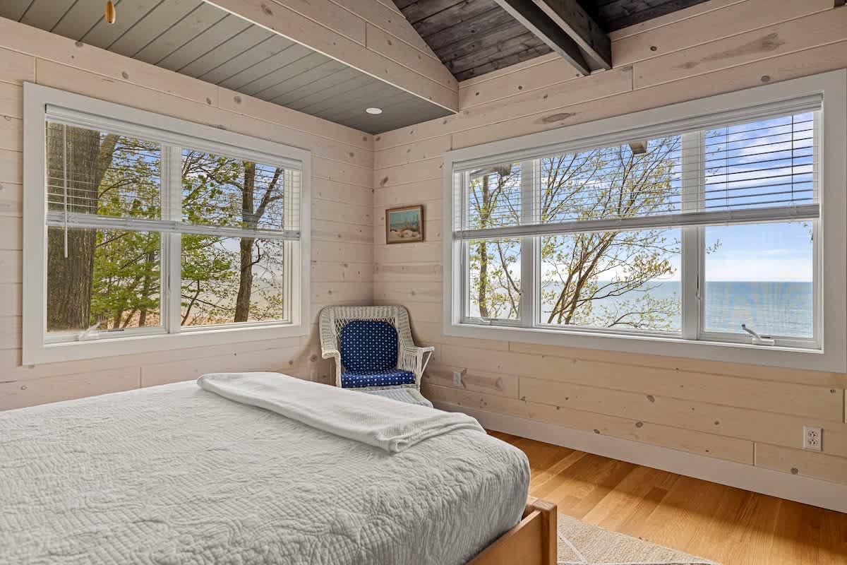 A bright bedroom with light wood paneled walls and a white bed, featuring two large windows offering views of trees and the ocean. A wicker chair with a blue cushion sits in the corner.