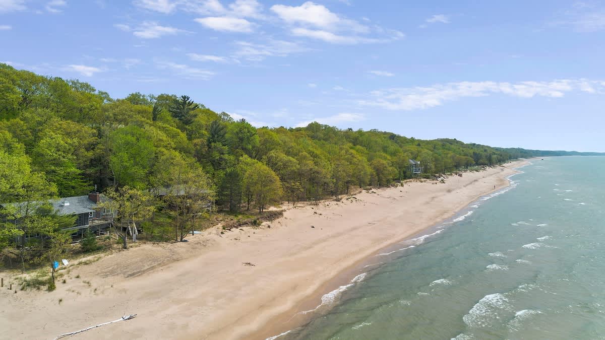 An aerial view showcasing a beachfront house nestled among lush green trees, with a wide sandy beach stretching along the turquoise ocean under a blue sky.