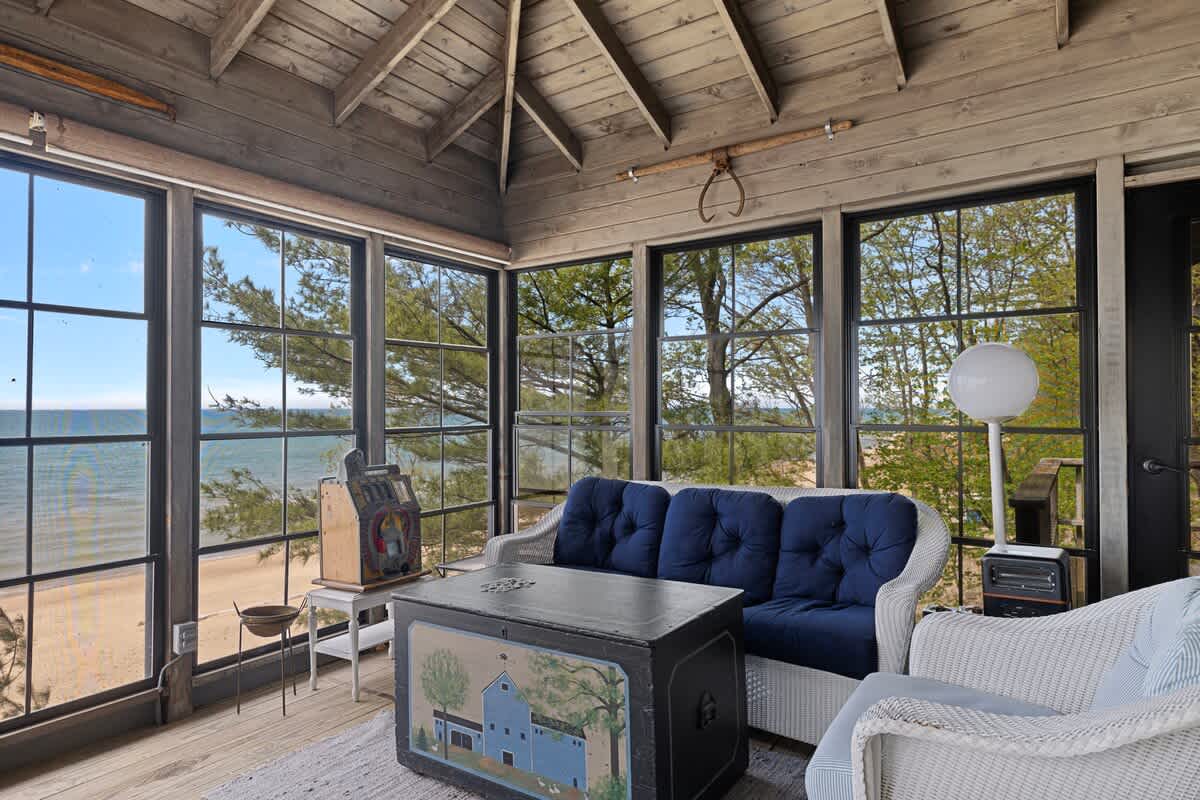 A sunroom living area with a white wicker sofa and blue cushions, facing large windows that overlook a sandy beach and the ocean. The room features a dark wooden vaulted ceiling and light wooden floors.