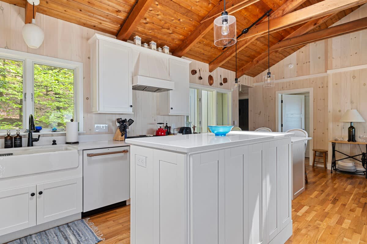 Modern white kitchen with a central island, a white farmhouse sink, and light wood floors under a vaulted wood ceiling. The space is well-lit with natural light from a window above the sink and features pendant lighting.