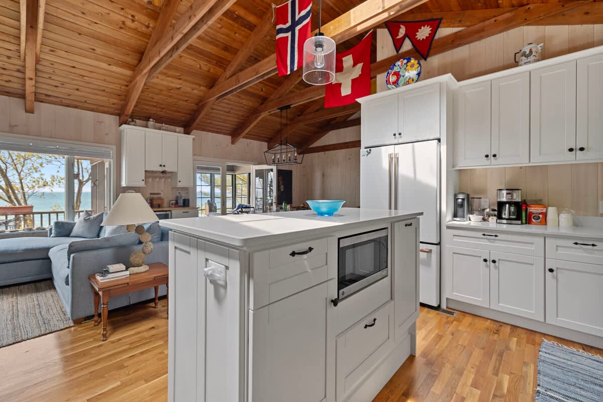 White kitchen island with a built-in microwave, set on light wood floors under a vaulted wood ceiling adorned with international flags. A light blue sofa is visible in the adjacent living area.