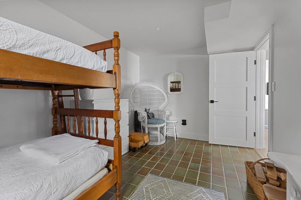 A children's bedroom featuring a wooden bunk bed with white bedding, green tiled flooring, and a white wicker chair in the corner, illuminated by recessed lighting.