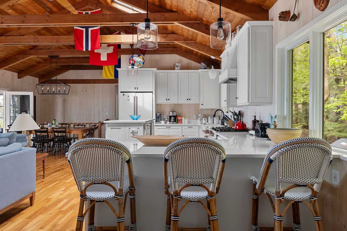 Modern white kitchen with a large island featuring three wicker bar stools, set against light wood floors and a vaulted wood ceiling. The kitchen is equipped with stainless steel appliances and offers a view of the surrounding greenery through large windows.