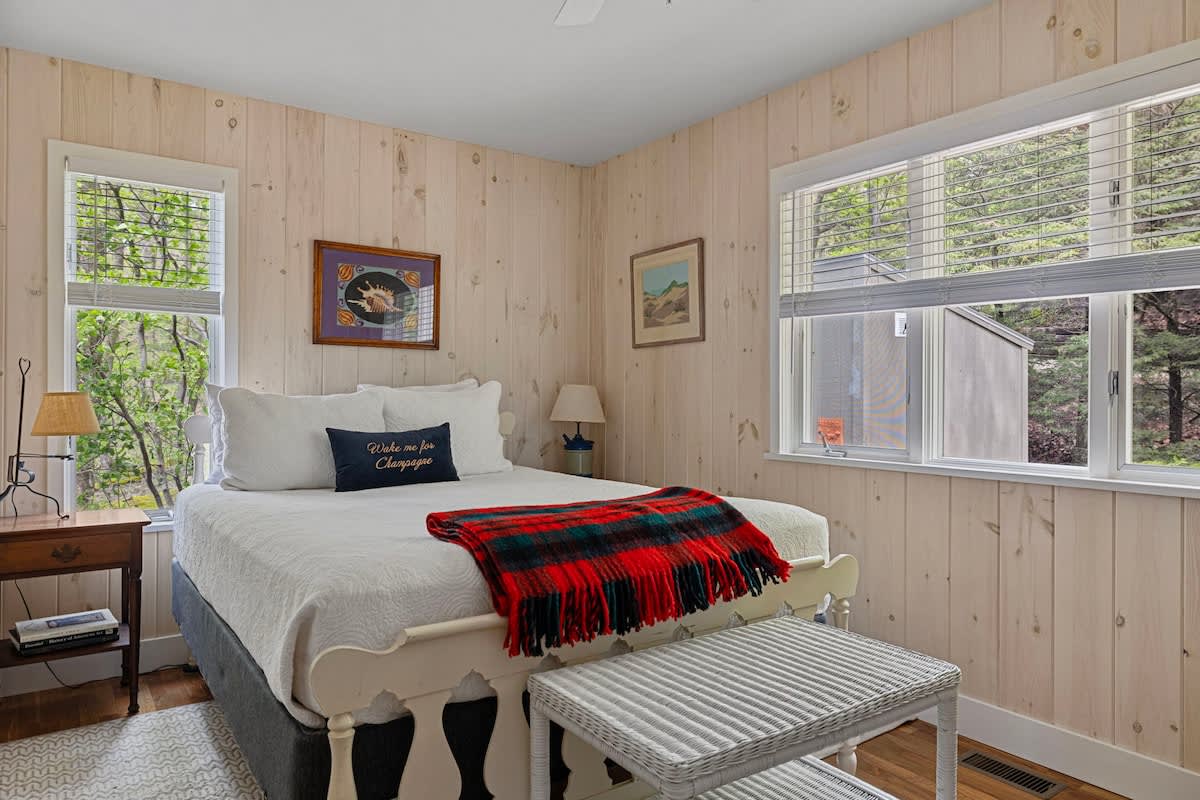 Cozy bedroom with light wood paneled walls, a white metal-frame queen bed with a red plaid throw, and two windows with blinds overlooking lush greenery. A white wicker bench sits at the foot of the bed.