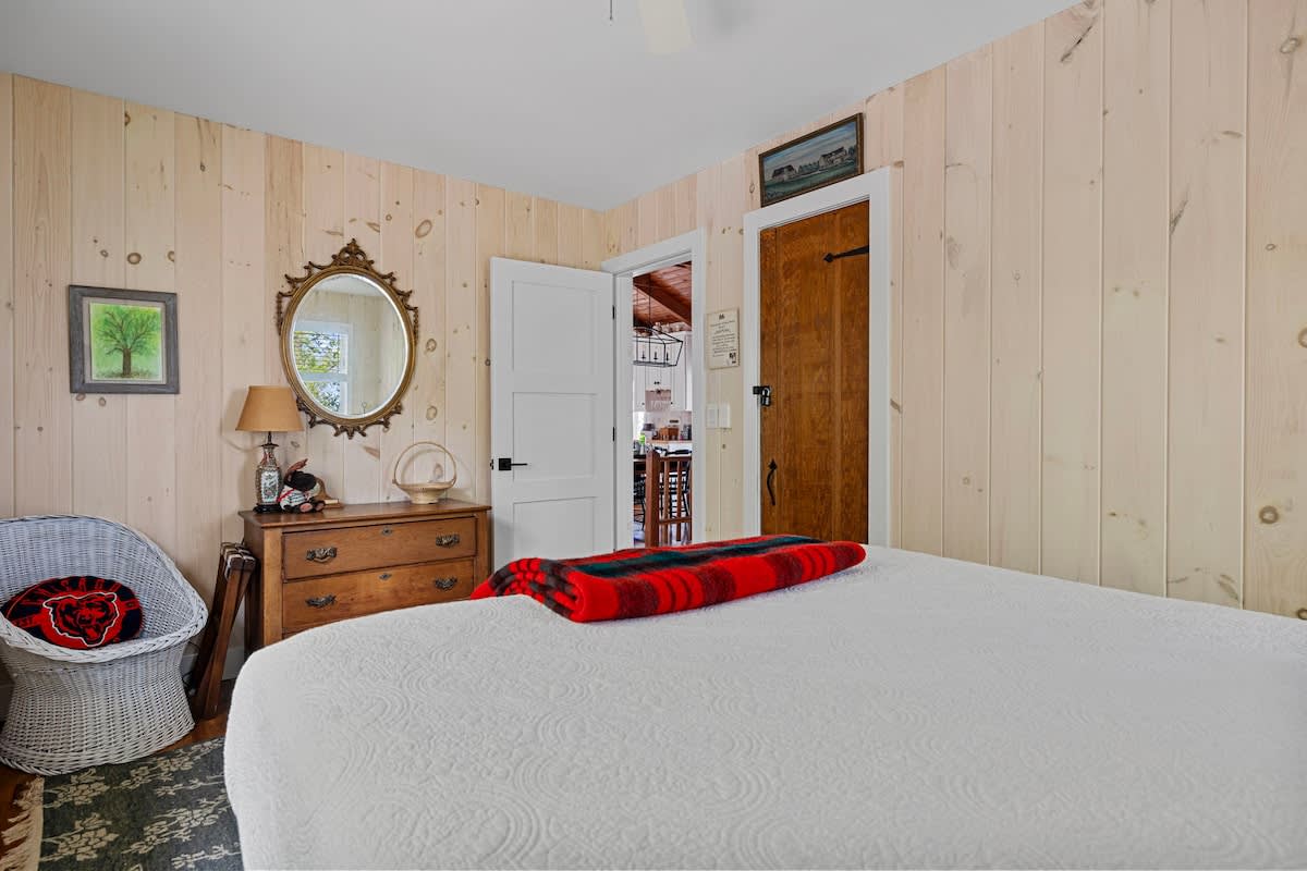 View of a bedroom with light wood paneled walls, showing a white bed with a red plaid throw and a vintage wooden dresser with an ornate mirror above it. A white wicker chair is in the corner.