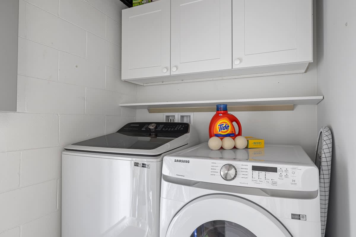 A functional laundry room equipped with a white Samsung top-load washer and front-load dryer, overhead white cabinets, and a shelf with laundry supplies.