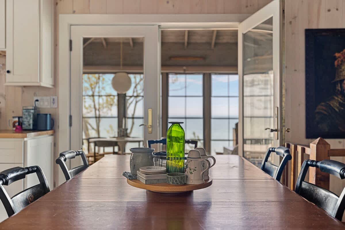 Close-up of a rustic wooden dining table adorned with a decorative centerpiece, with black chairs surrounding it. Large glass French doors in the background offer a glimpse of an outdoor area and a lake view.