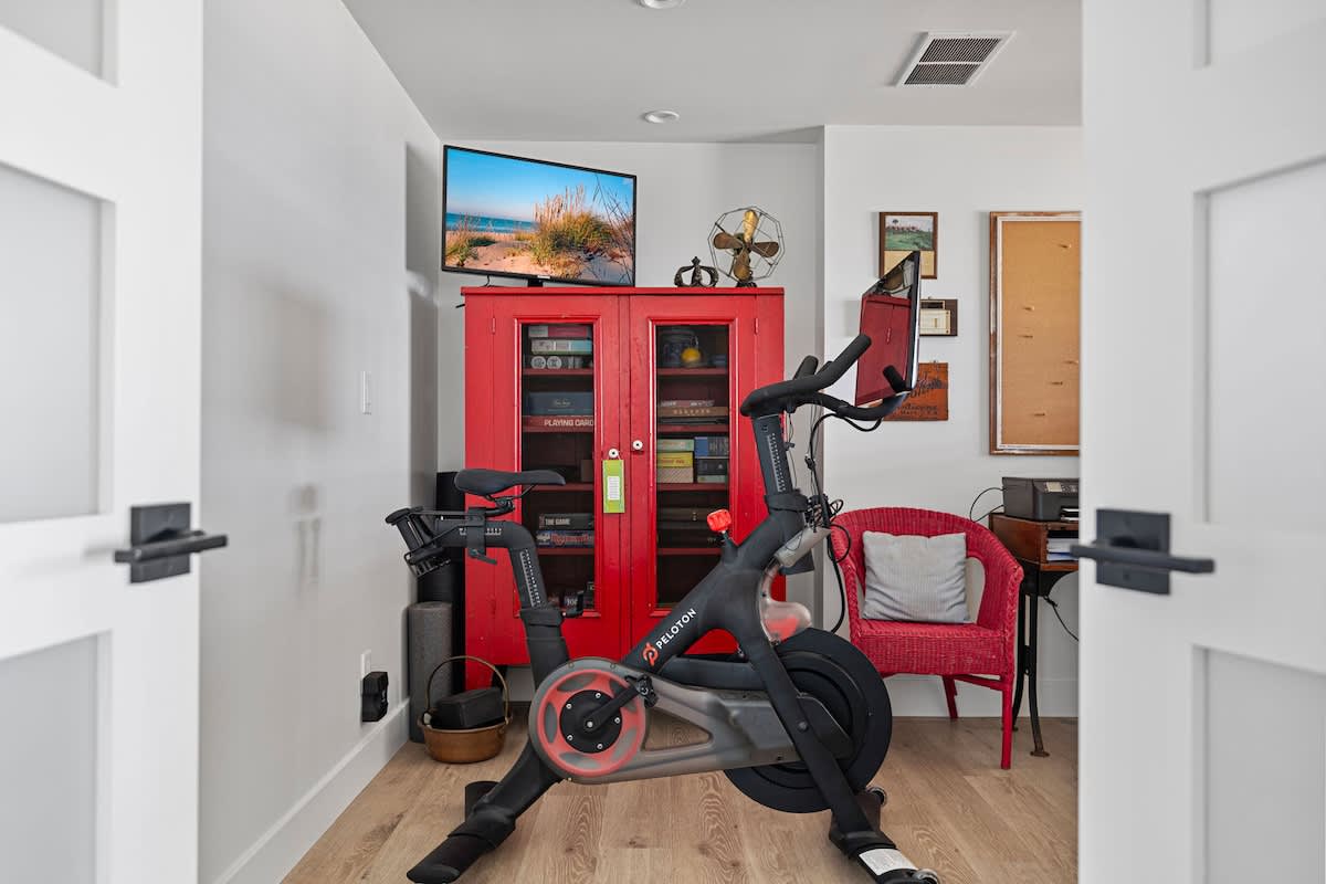A bright recreation area featuring a Peloton bike, a red storage cabinet with a TV on top, and a small red wicker chair, all on light wood-look flooring.