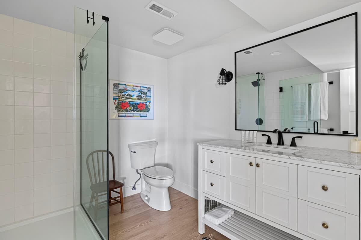 Modern bathroom with white walls and light wood flooring, featuring a white vanity with a marble top and black fixtures, and a glass-enclosed walk-in shower. A large black-framed mirror hangs above the vanity.