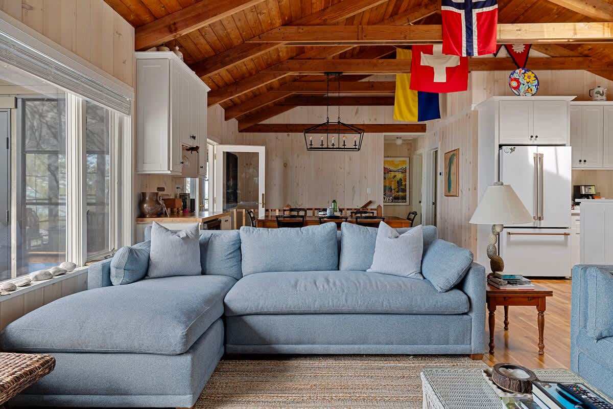 Expansive living room with a light blue sectional sofa and vaulted wood-paneled ceiling, flowing into an open-concept kitchen and dining area, adorned with nautical flags.