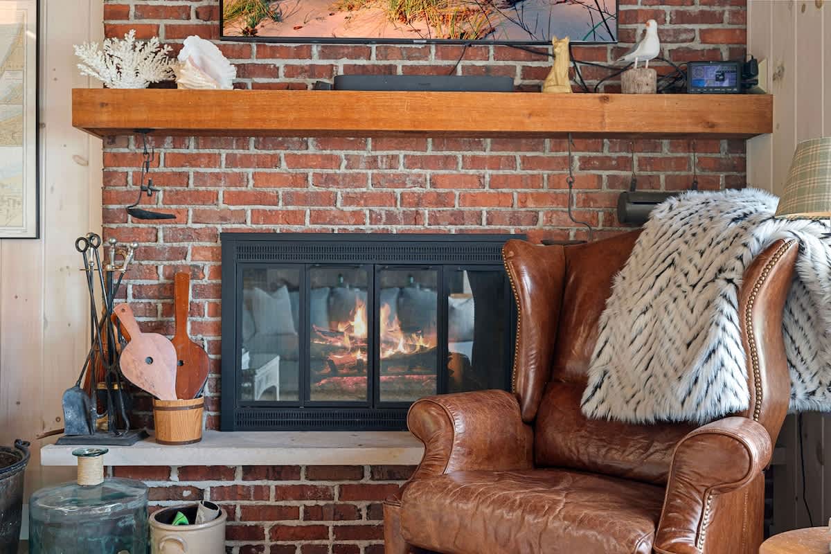 Close-up of a rustic brick fireplace with a roaring fire, a wooden mantel adorned with decorative items, and a cozy leather armchair with a faux fur throw.