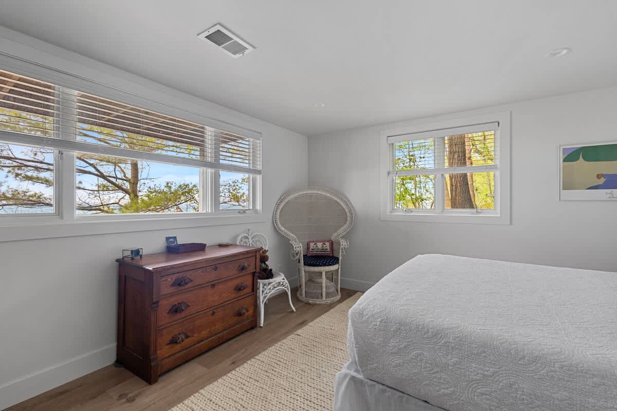 White-walled bedroom with light wood flooring, showcasing a vintage wooden dresser and a distinctive white wicker peacock chair. Large windows with blinds offer views of trees and distant water.