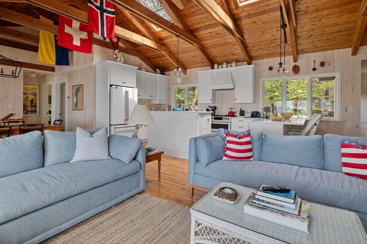 Open-concept living area featuring two light blue sofas with patriotic throw pillows, light wood floors, and a vaulted wood-paneled ceiling adorned with international flags. A modern white kitchen with an island is visible in the background.
