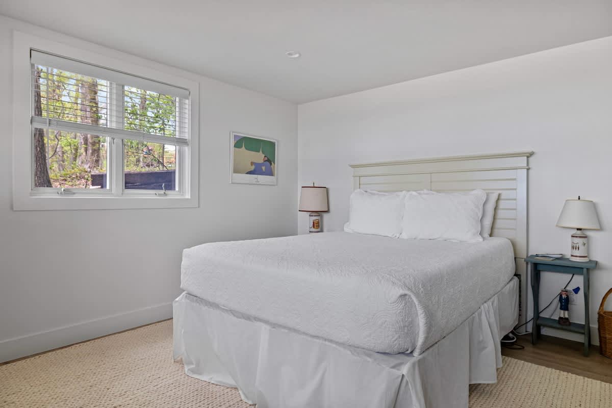 Bright bedroom with white walls and light wood flooring, featuring a white queen bed with a simple headboard and a large window with blinds offering a view of trees. A small blue nightstand with a lamp is beside the bed.