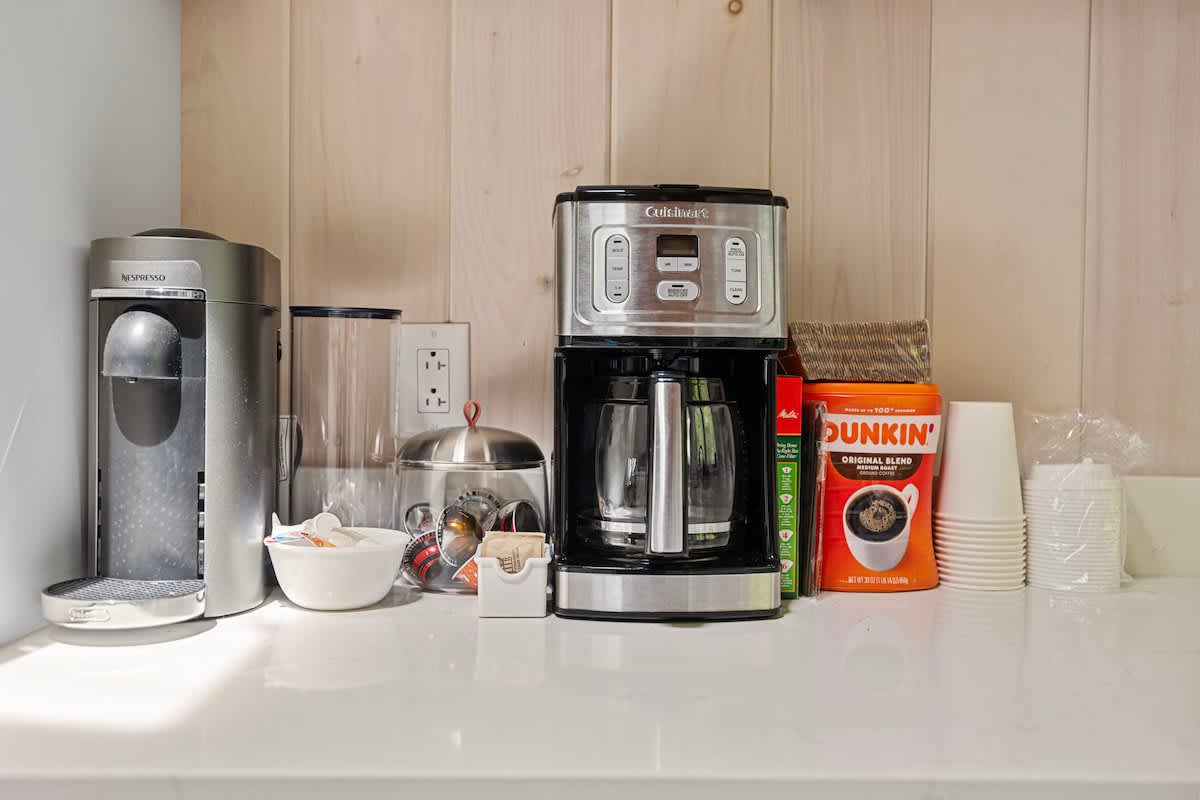 Dedicated coffee station on a white countertop, featuring a Nespresso machine, a drip coffee maker, and various coffee supplies. The area is backed by light wood paneling.