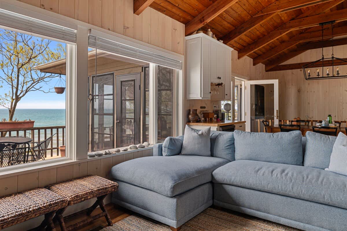 Bright living room featuring a light blue sectional sofa, woven ottomans, and large windows providing a stunning view of the lake and an outdoor deck.