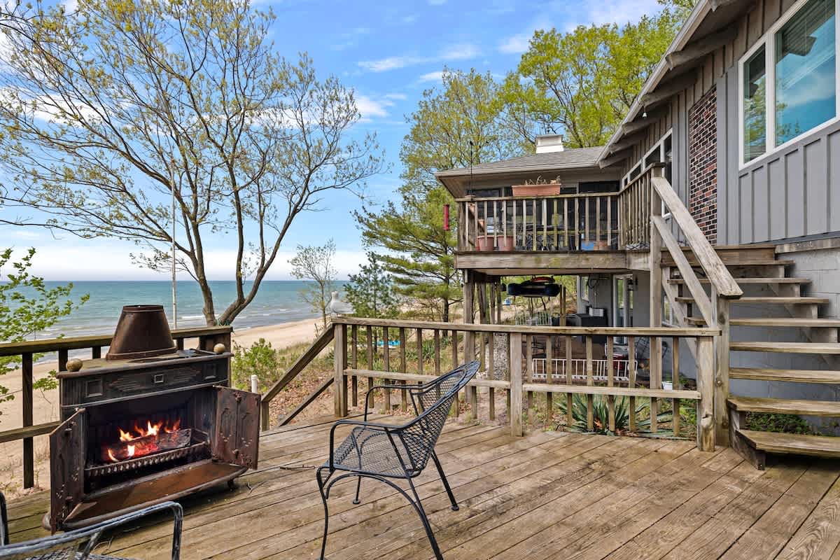 Spacious wooden deck overlooking a sandy beach and the lake, featuring an outdoor fireplace, metal chairs, and stairs leading down to the lower level and beach access.