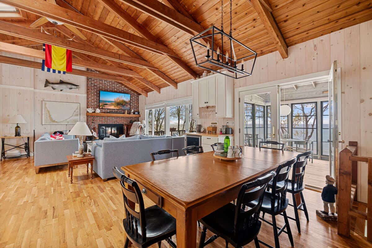 Spacious dining area with a large wooden table and black chairs, set on light hardwood floors under a vaulted wood-paneled ceiling. A brick fireplace with a TV is visible in the adjacent living room, and large glass doors lead to an outdoor space.